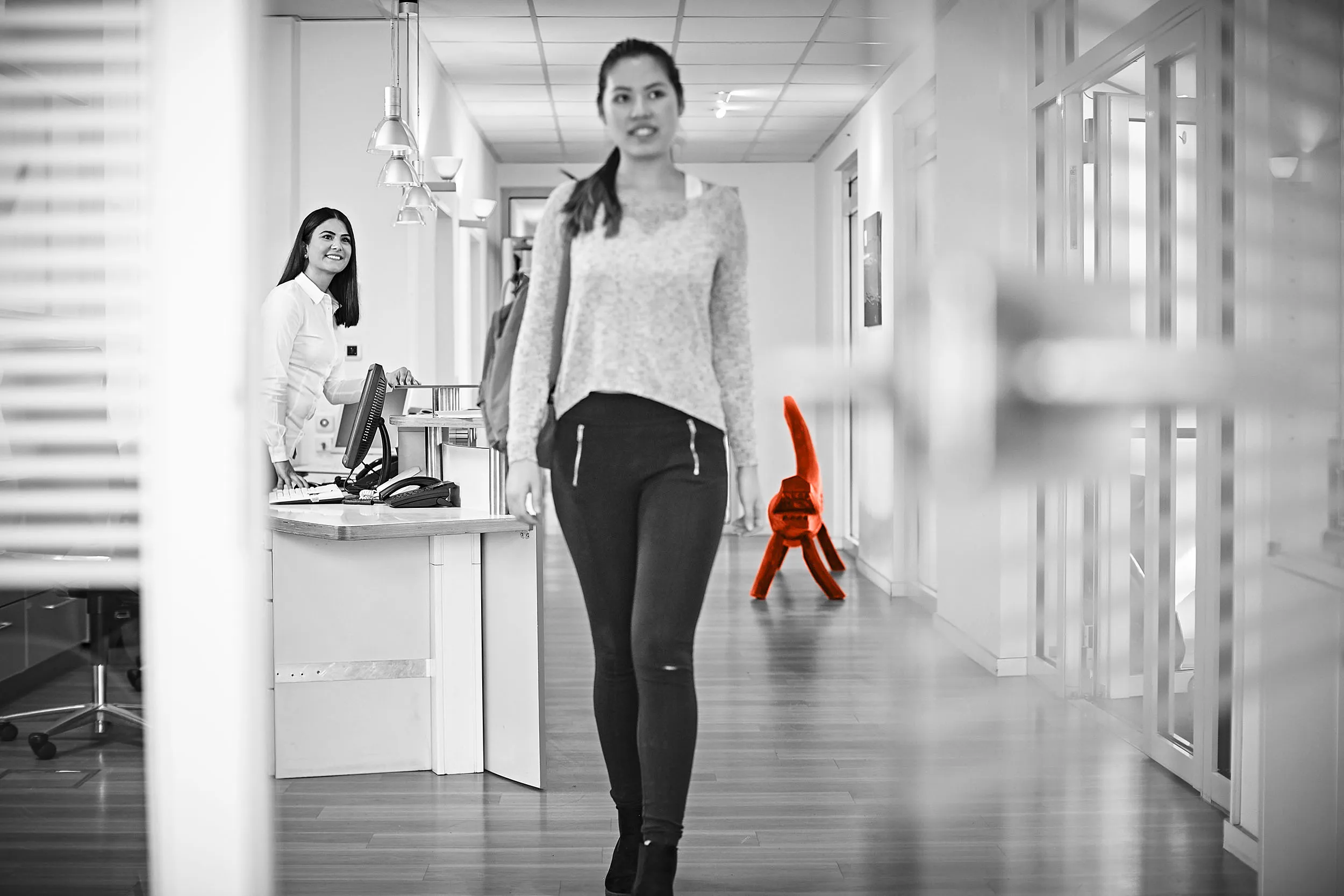 Wir helfen Ihnen gerne in unserer kieferorthopädischen Praxis weiter! Patientin geht den Flur entlang und im Hintergrund schaut die Mitarbeiterin am Empfang ihr nach.